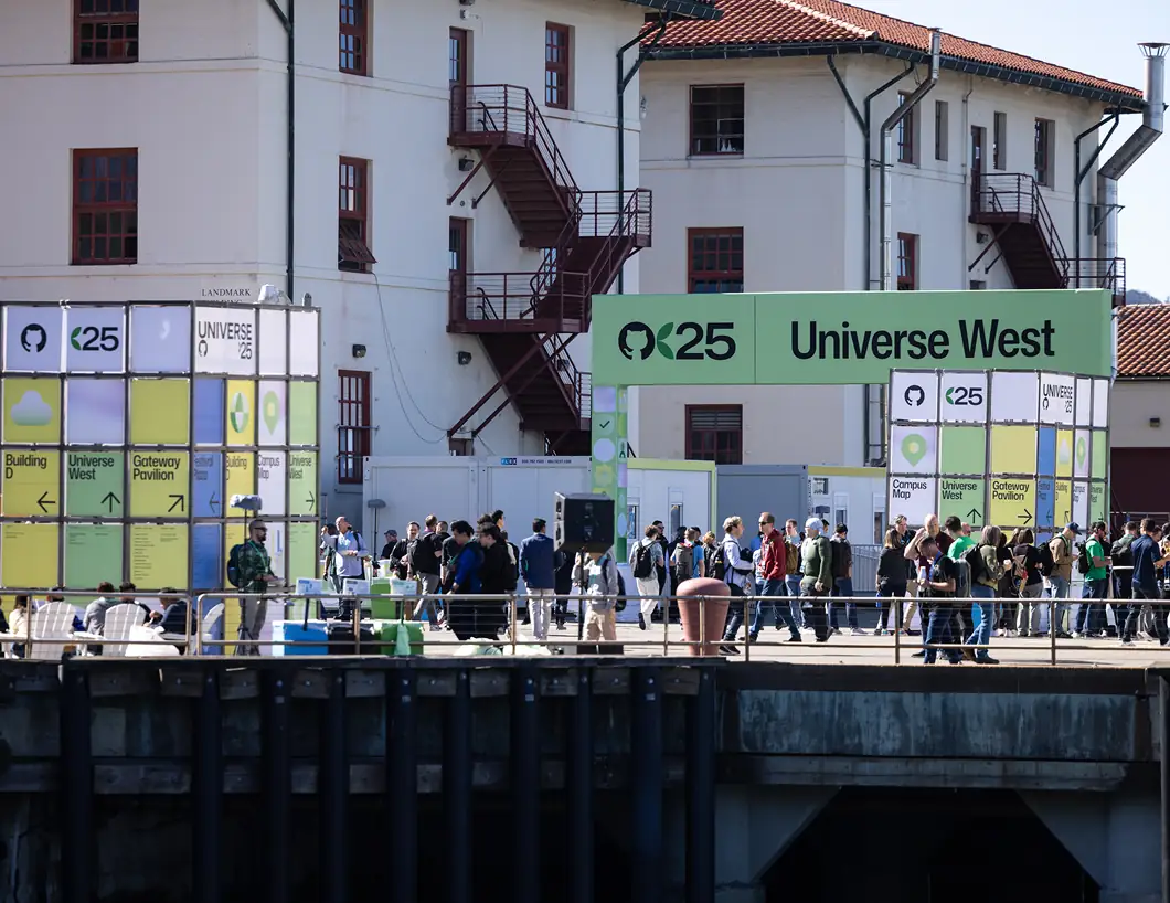 An outdoor view of the GitHub Universe conference entrance. A large green sign overhead reads "Universe West" alongside the GitHub logo and the number "25." Large, colorful, modular cubes display event maps and schedules, while attendees walk around a sunlit courtyard near a historic white building.