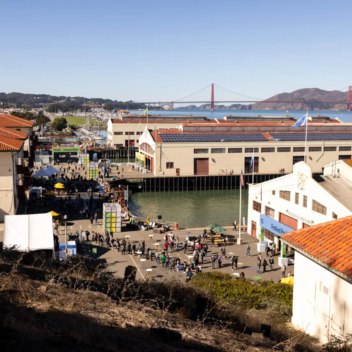 A wide daytime view of the GitHub Universe event at San Francisco’s waterfront, with attendees walking between pier buildings and the Golden Gate Bridge visible in the background.