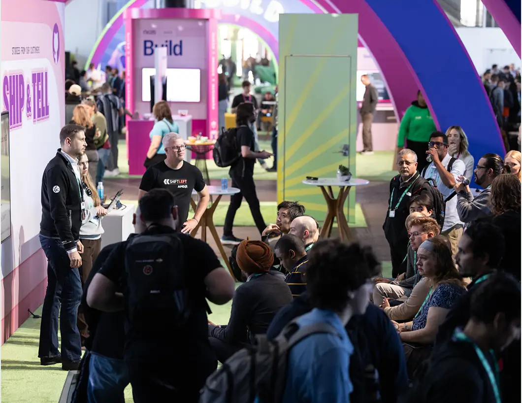 A bustling indoor scene at the conference. Attendees are gathered in a brightly lit, modern hall with pink and blue architectural arches. Some people are standing and talking near high-top tables, while others are seated, listening to a presentation or "Ship & Tell" session.