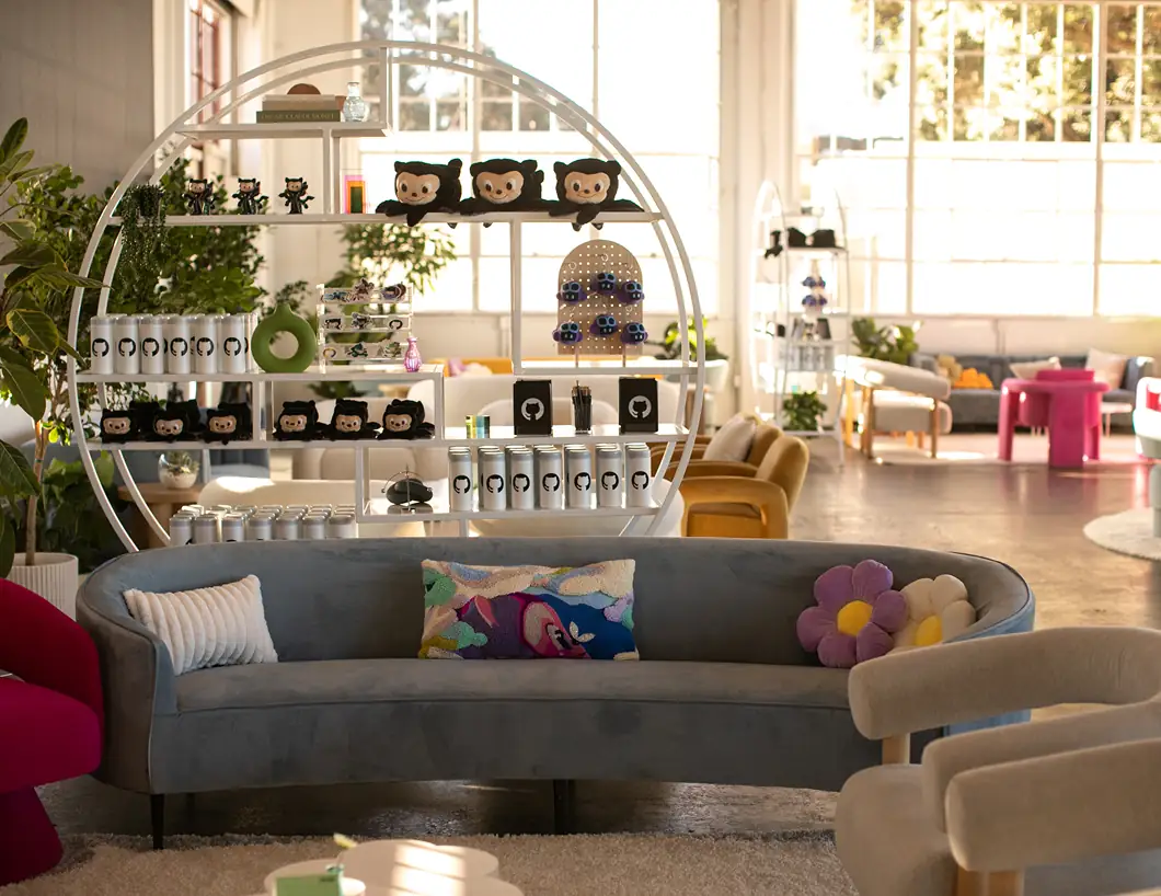 A stylishly decorated lounge area featuring a grey velvet sofa with colorful flower-shaped pillows. Behind the sofa is a large, circular white shelving unit filled with GitHub merchandise, including "Mona the Octocat" plushies and branded water cans. Large windows in the background fill the room with natural light.